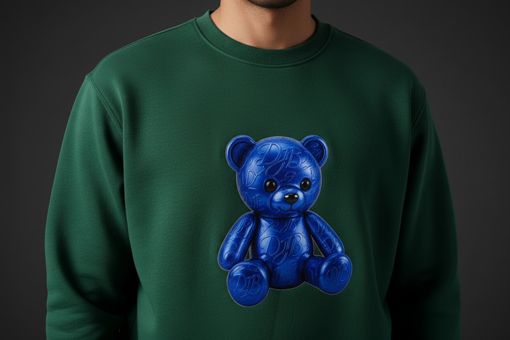 Close-up of the green DJB Teddy heavyweight crewneck showing the blue teddy print, fleece texture, stitching, and ribbed cuff